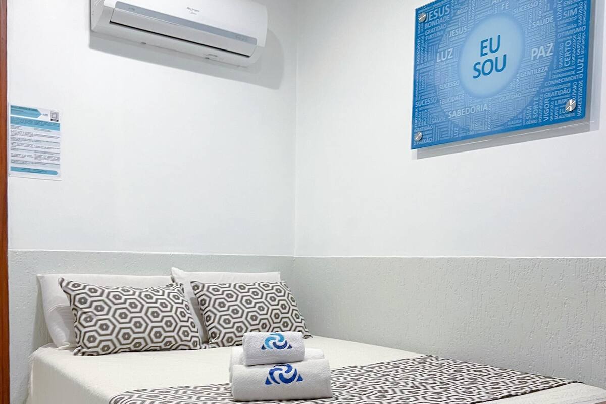 A hotel room with a bed, patterned pillows, a blue wall with 'EU SOU' text, and an air conditioning unit.