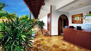 Zona de recepción de un hotel con suelo de baldosas, mostrador de madera y una gran planta en maceta.