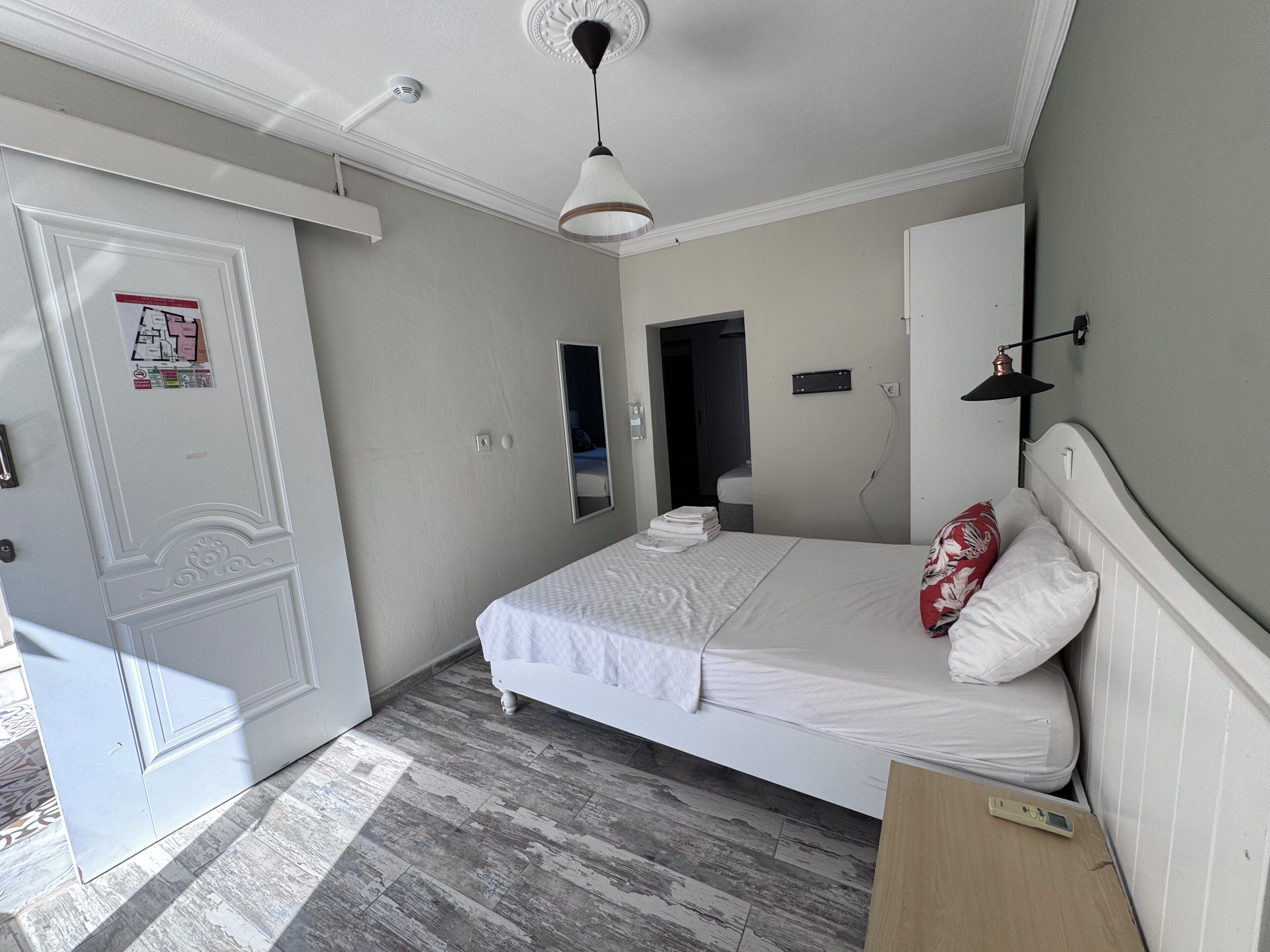 A bedroom with a bed, a bedside table, a mirror, a door, and a light fixture.