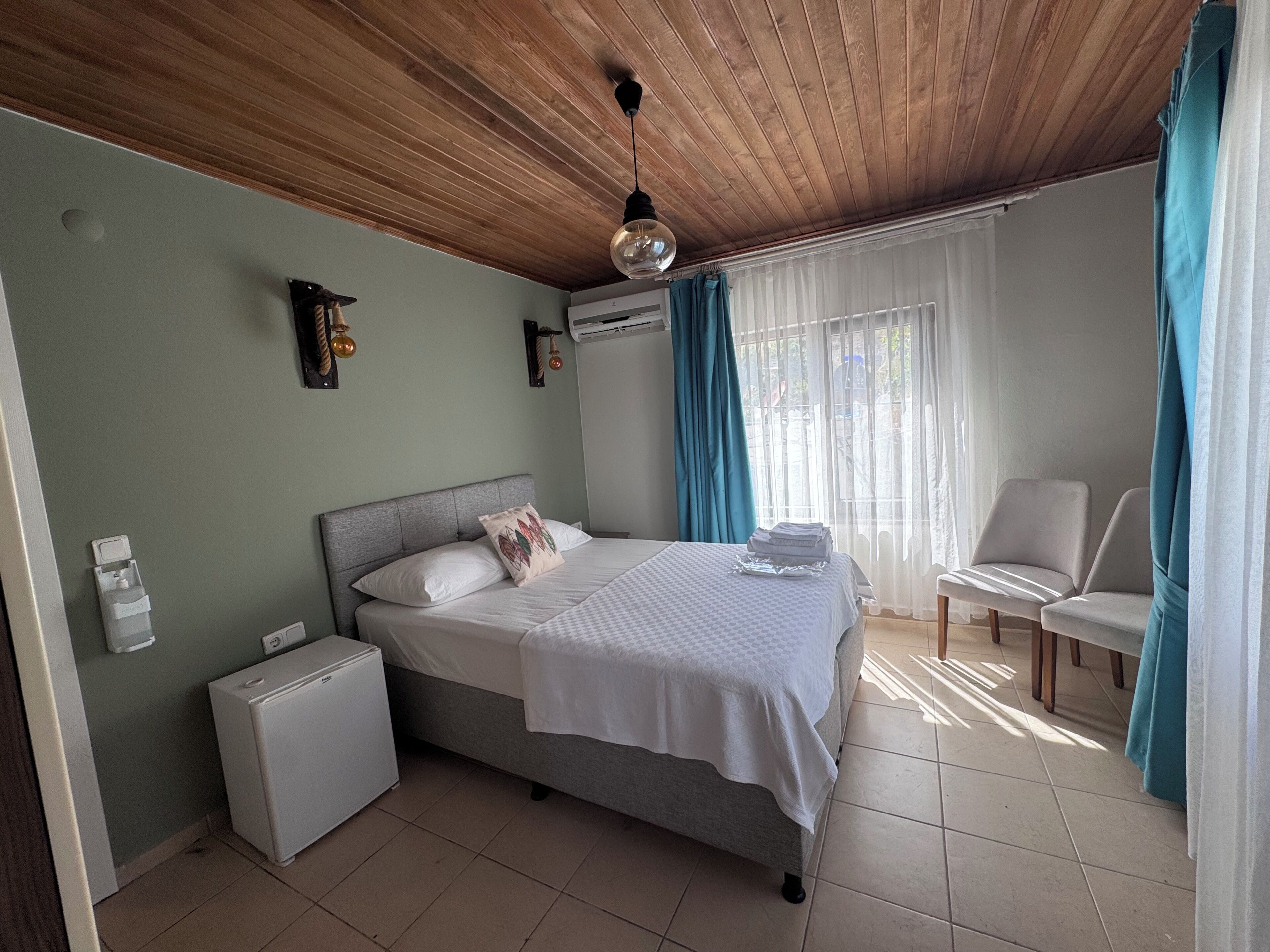 A bedroom with a bed, a small white refrigerator, a chair, and a window with curtains.