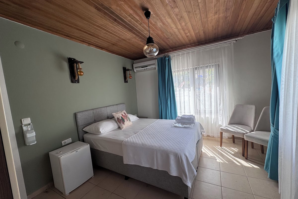 A bedroom with a bed, a small white refrigerator, a chair, and a window with curtains.