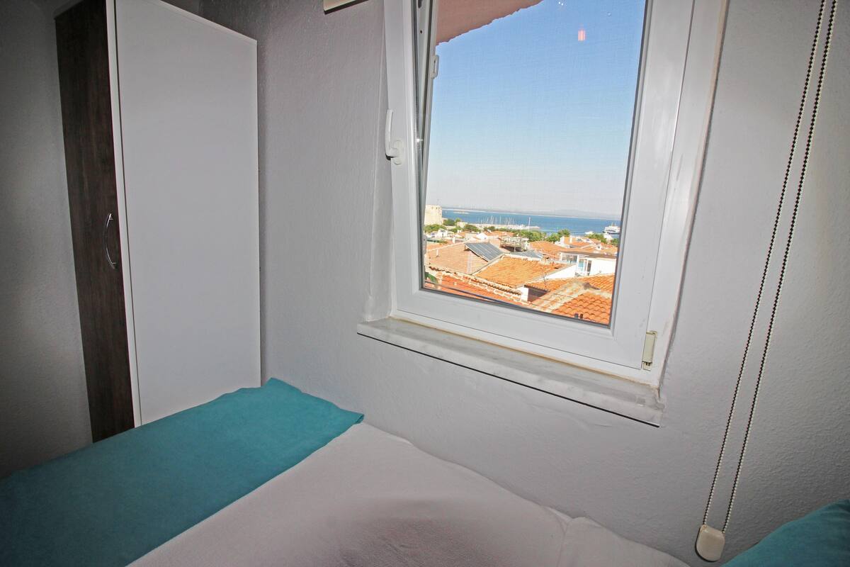 A bedroom with a window view of a coastal town, a bed, and a wardrobe.