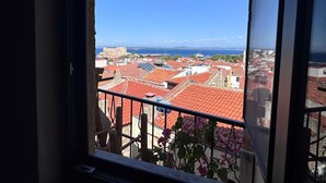 Vista desde una ventana sobre un pueblo costero con edificios de techo rojo y un mar lejano.