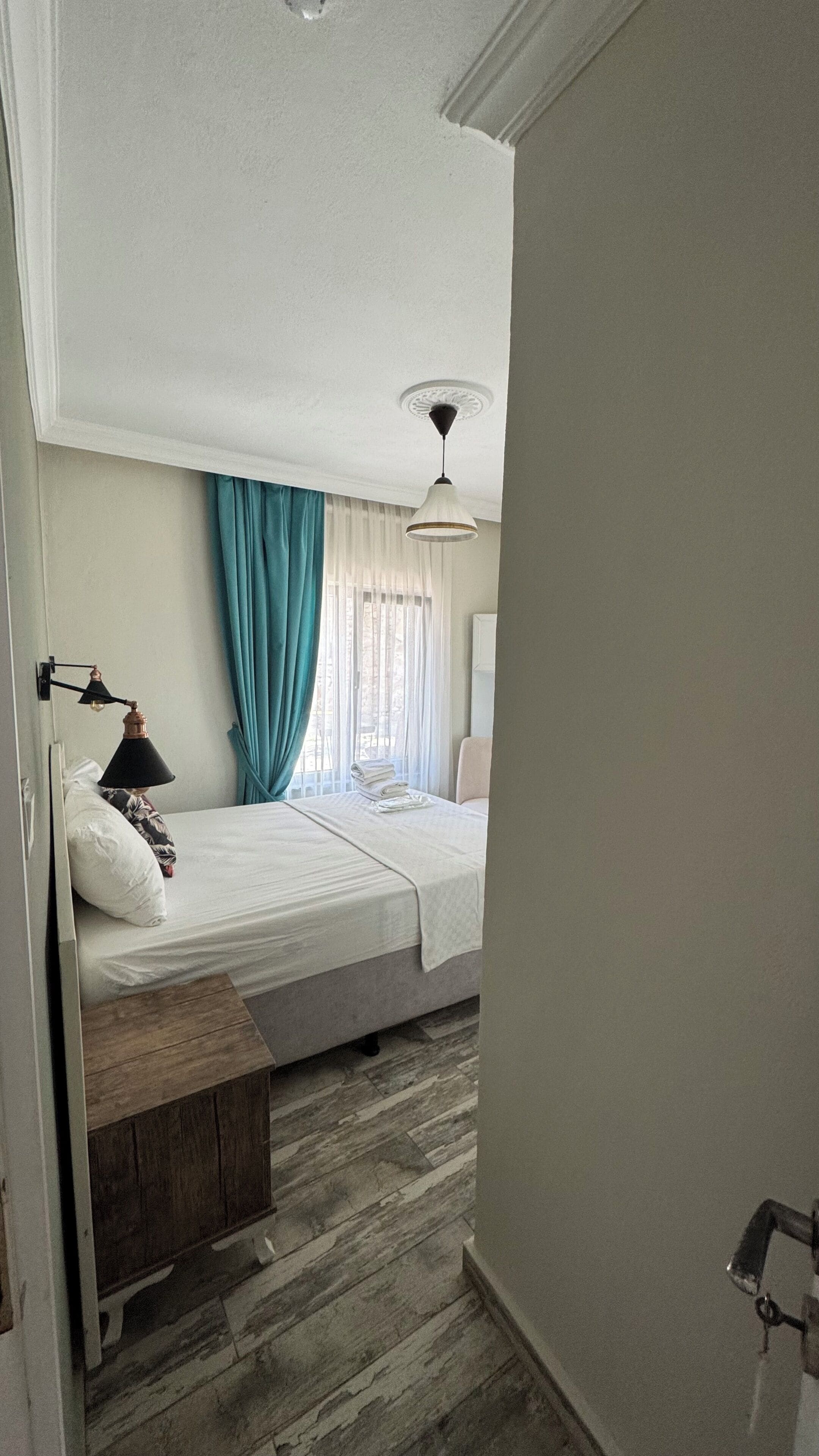 A hotel room with a bed, a bedside table, a lamp, a ceiling light, and a window with curtains.