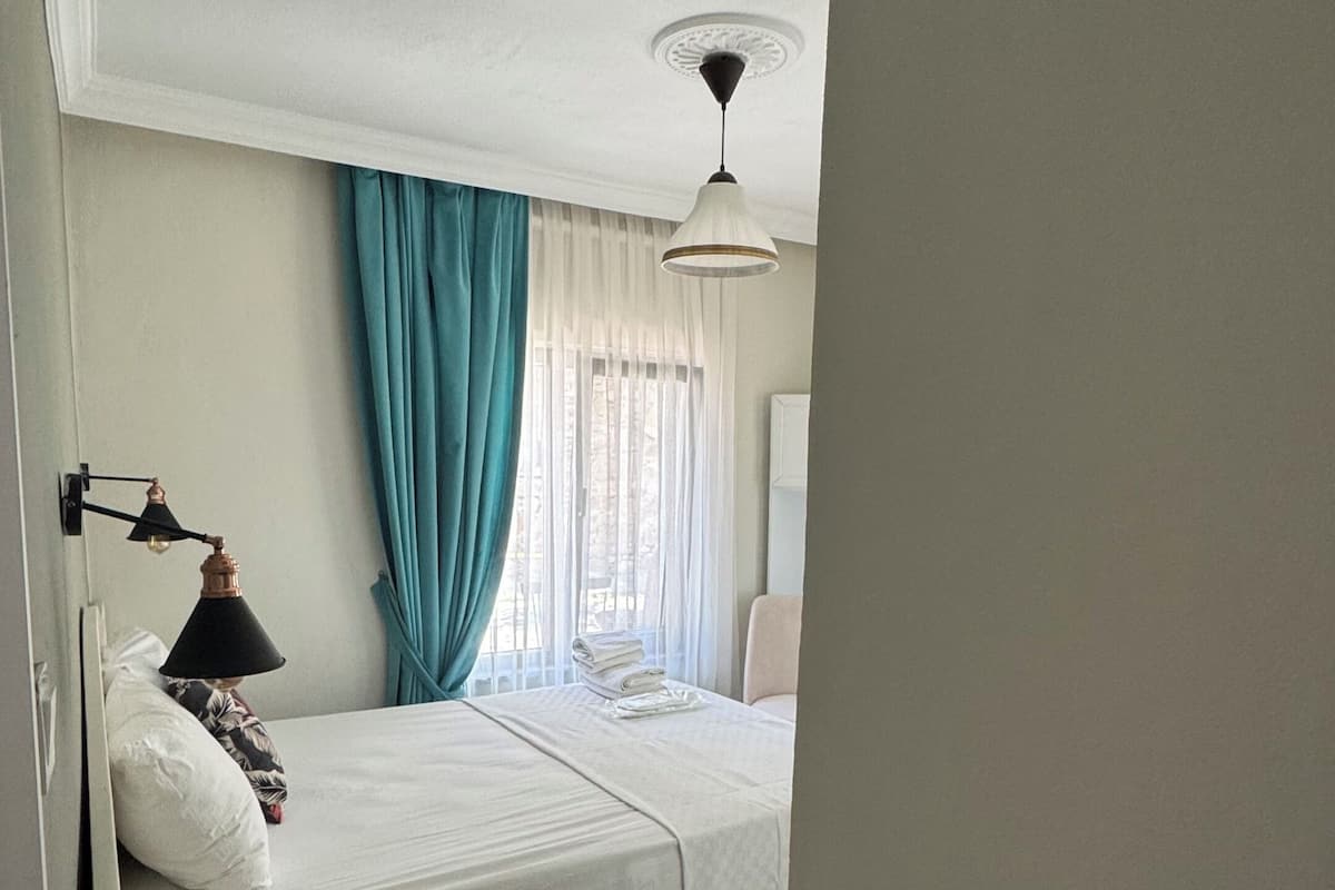 A hotel room with a bed, a bedside table, a lamp, a ceiling light, and a window with curtains.