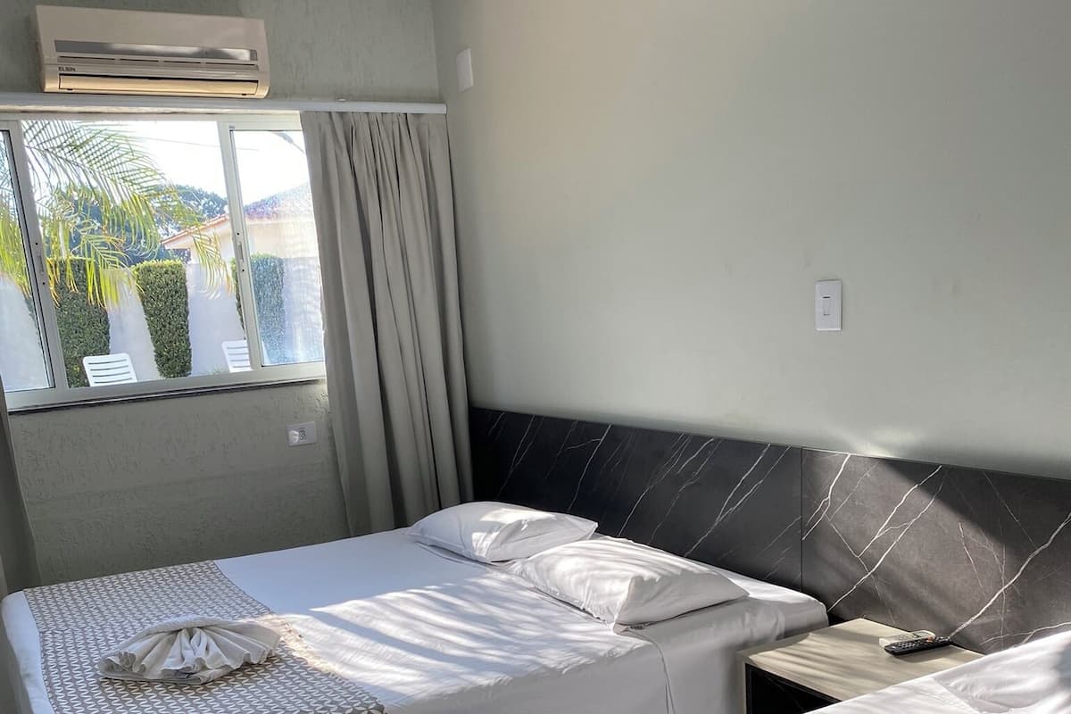A hotel room with two beds, a marble headboard, an air conditioning unit, a window with curtains, and a view of greenery outside.