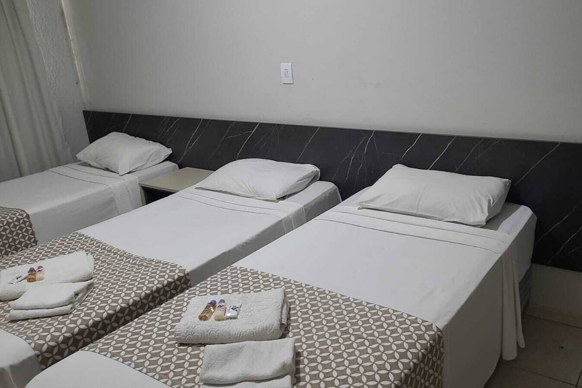 A hotel room with three single beds, each with a patterned bedspread and a white pillow. There are towels and toiletries on the beds.