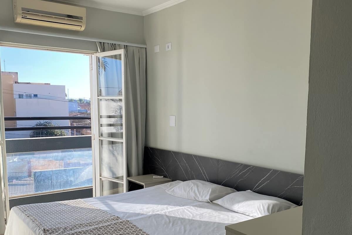 A hotel room with a bed, a window with curtains, an air conditioning unit, and a view of buildings outside.