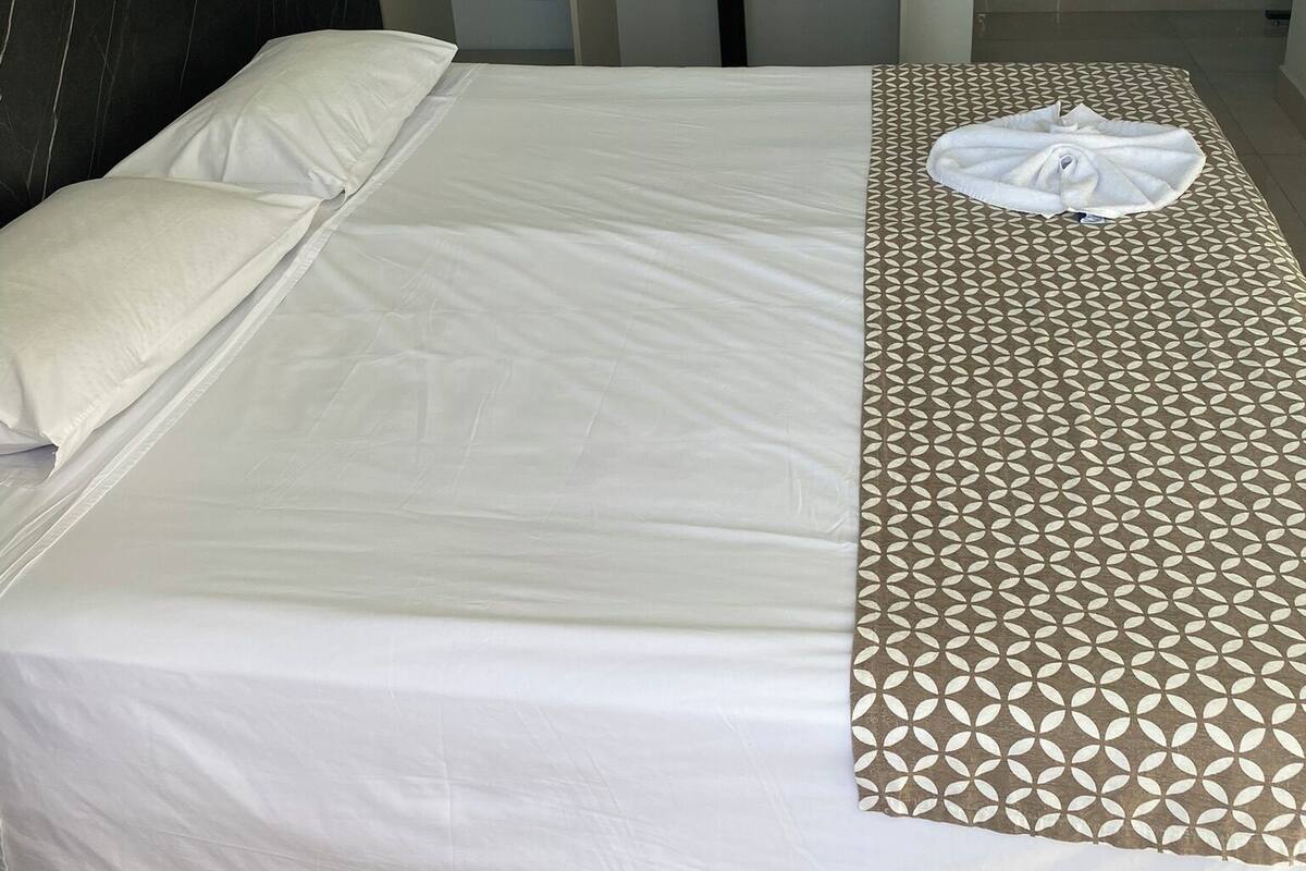 A neatly made bed with a patterned bedspread and pillows, a small desk with a chair, and a grey pillow on the shelf.