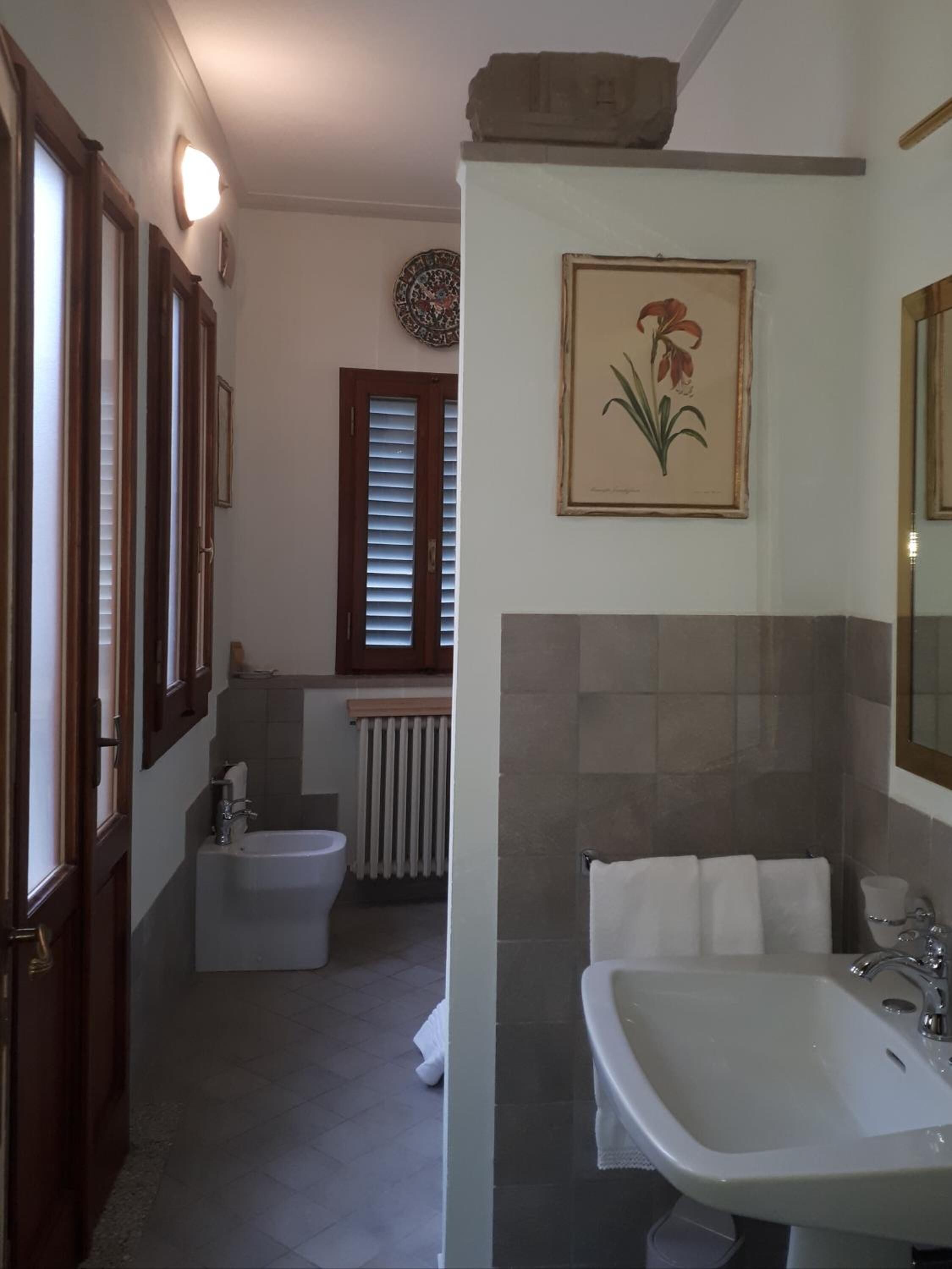 Casa de banho com lavatório, bidé e sanita. Tem uma janela com persianas e um radiador.