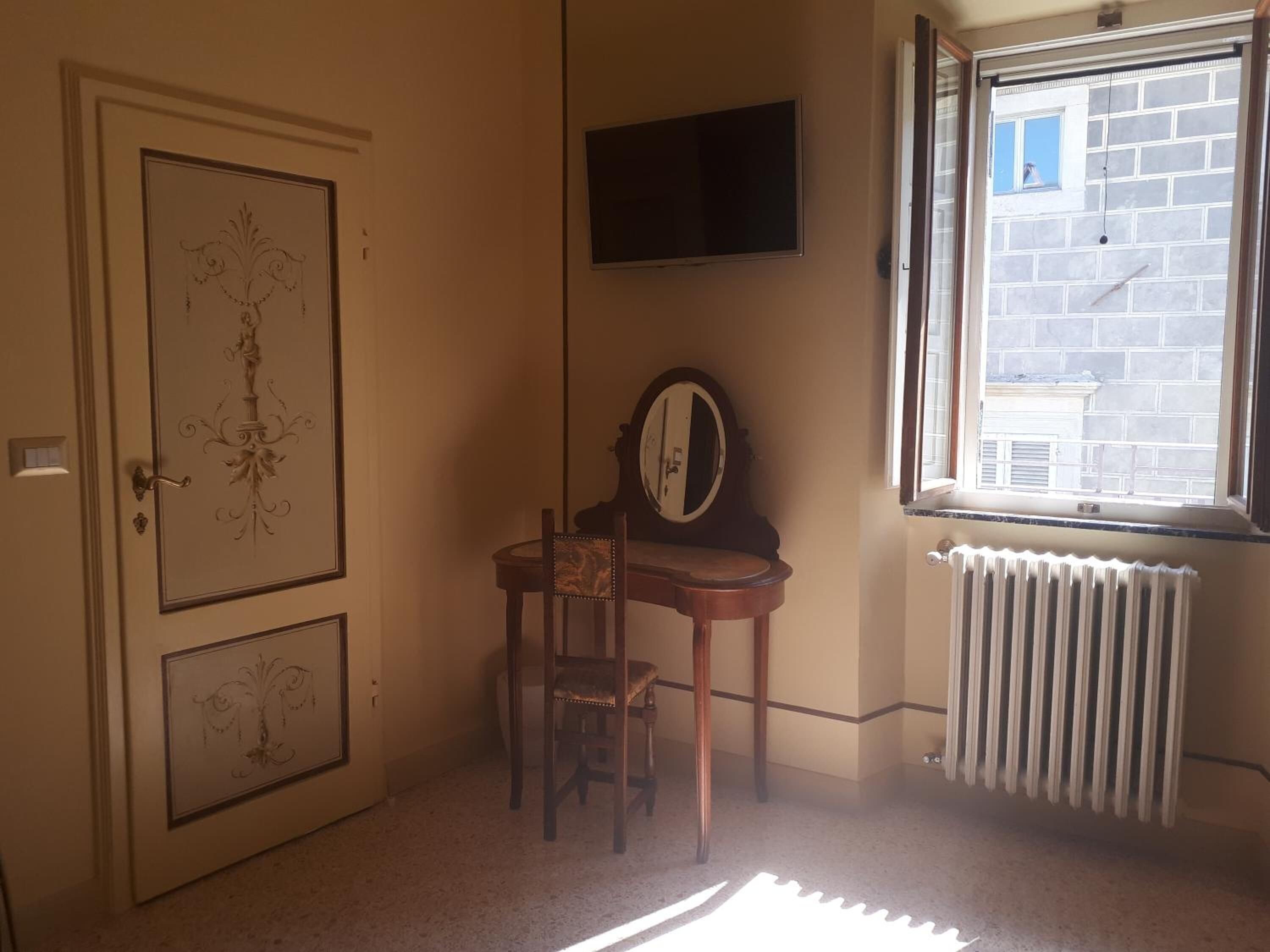 A room with a door featuring a decorative design, a small table with a chair, a mirror, a television mounted on the wall, and a window with a view of a brick building.