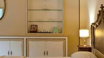 Et hotelværelse med seng, et natbord med lampe, en kommode med spejl og en hylde med dekorative genstande.