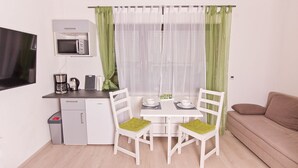 A compact kitchen with a microwave, coffee maker, and a small trash bin. There is a dining table with two chairs, a sofa, and sheer curtains. - Ferienwohnung BECKENDORF 3 (Losheim am See)