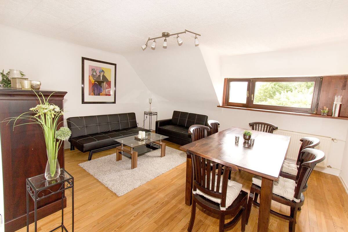 A living room with a black sofa, a glass coffee table, a wooden dining table with chairs, a white rug, and a window with a view of greenery.