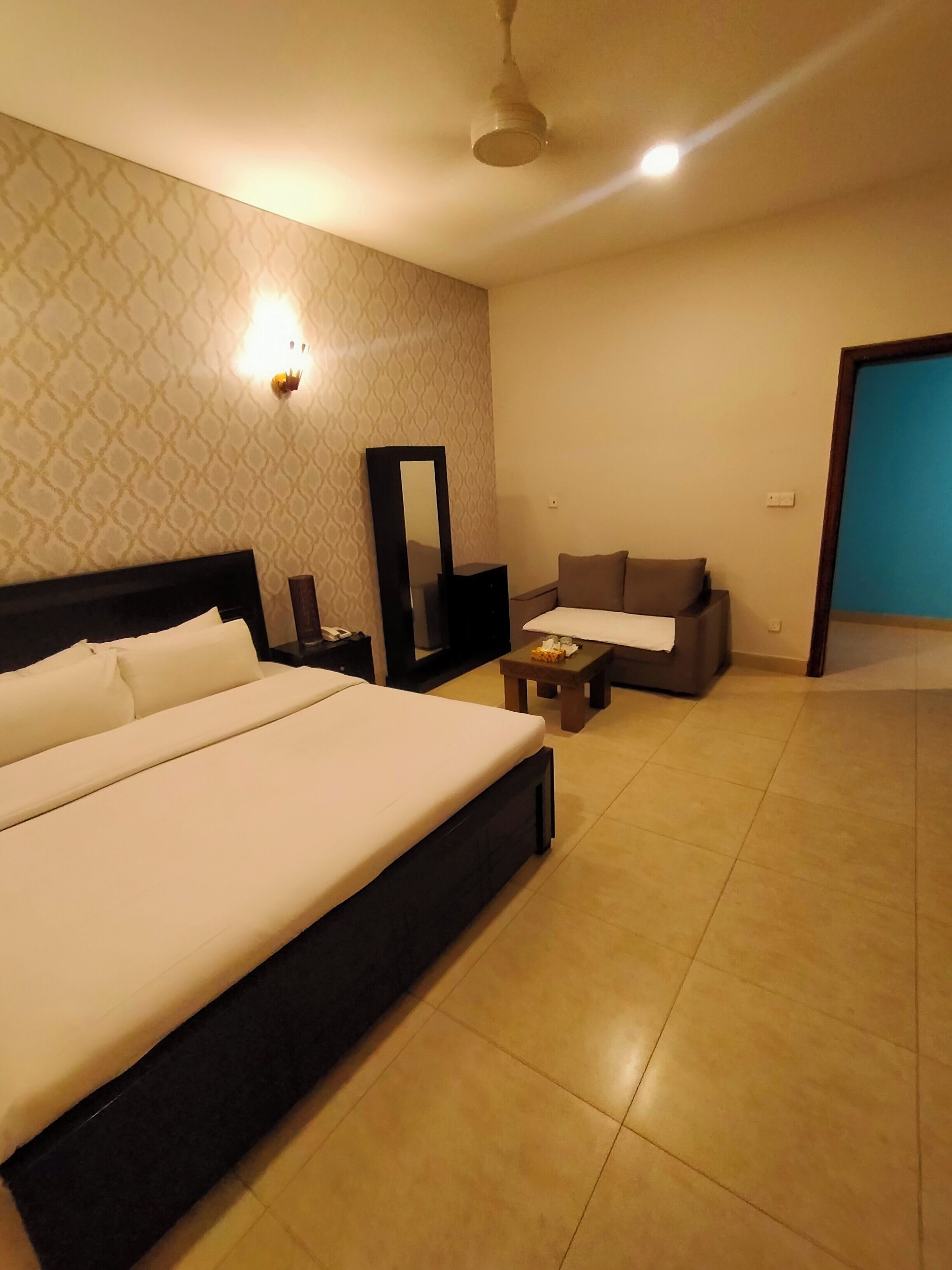 Habitación de hotel con una cama grande, un pequeño sofá, una mesita de noche, un espejo y un ventilador de techo.