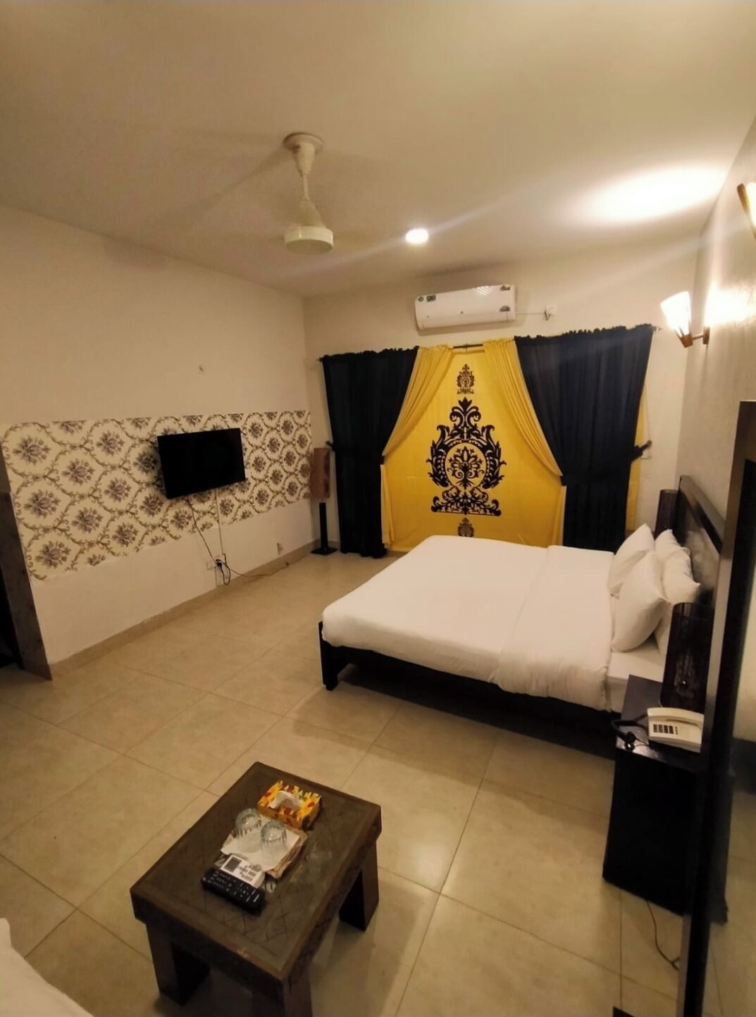 A hotel room with a bed, a small table, a flat-screen TV mounted on the wall, and an air conditioning unit.