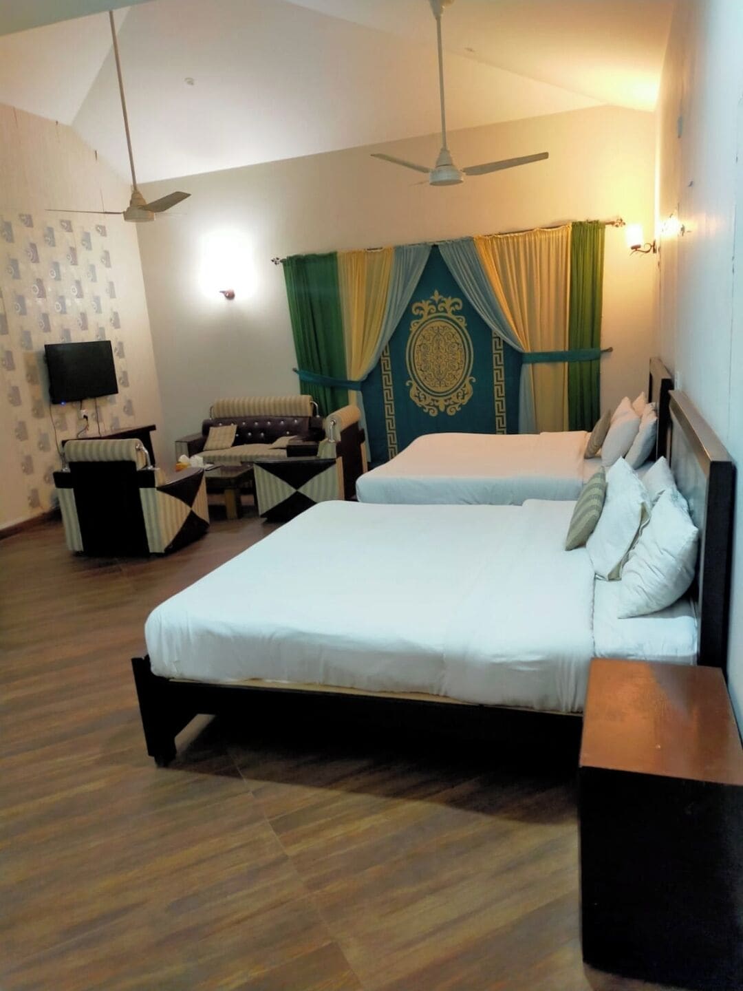 A hotel room with two beds, a sofa, a TV, and a ceiling fan.