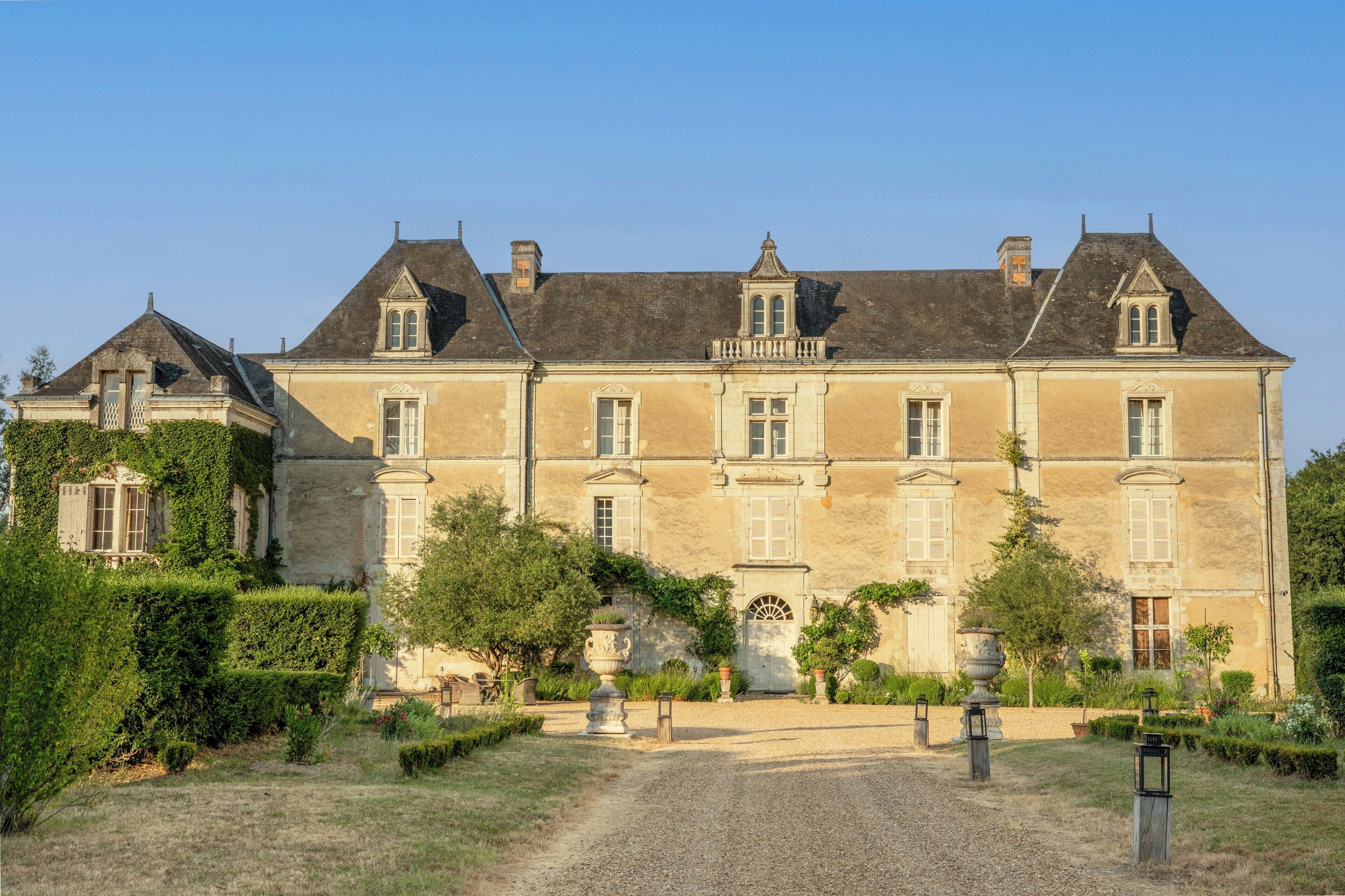 A large, historic building with multiple windows and a symmetrical facade, surrounded by a well-maintained garden with gravel paths.