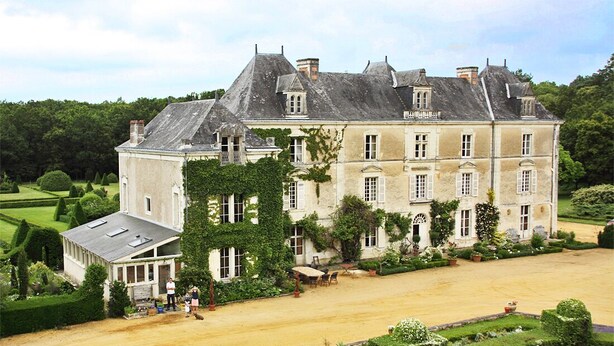 Un grand bâtiment historique à la façade symétrique, doté de nombreuses cheminées et agrémenté d’un jardin bien entretenu avec des haies et une pelouse.