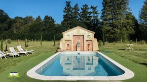 Une piscine avec une terrasse carrelée, un petit bâtiment avec une entrée carrelée et une pelouse agrémentée de chaises longues.
