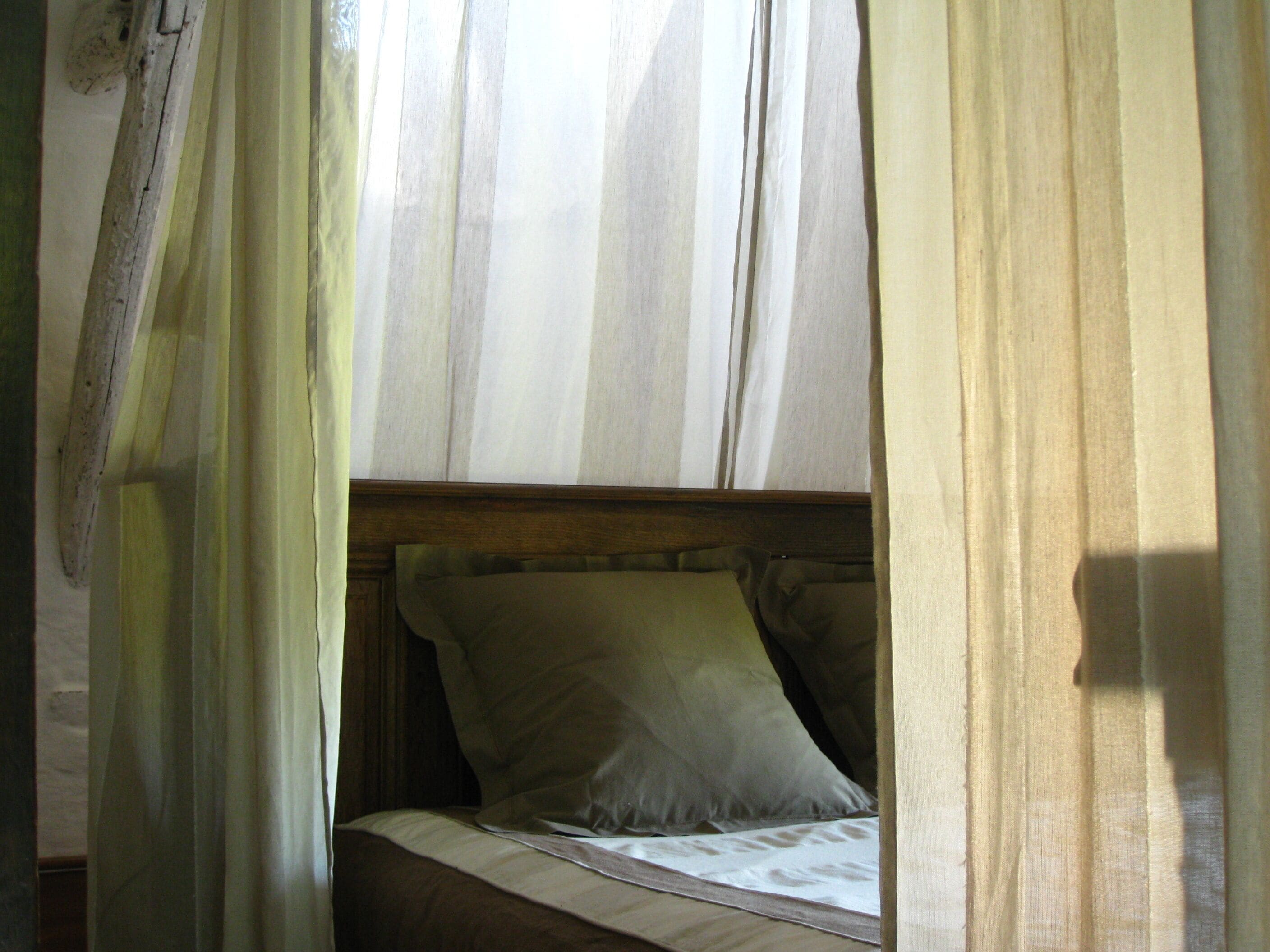 A single bed with a wooden headboard and white bedding, partially obscured by sheer curtains.