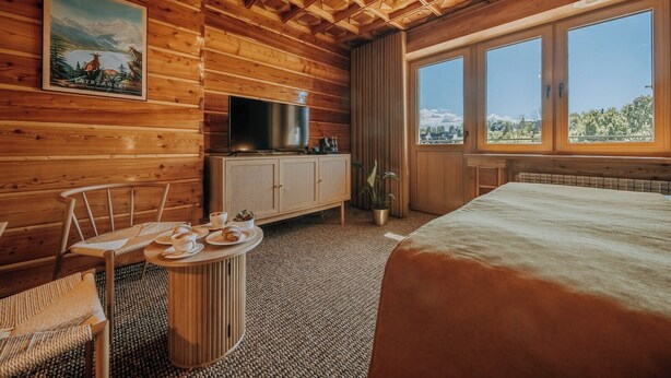 A wooden cabin room with a bed, a television, a dining table with plates and cups, and a window with a view of the mountains. - Dom Wypoczynkowy U Staszla (Szaflary)