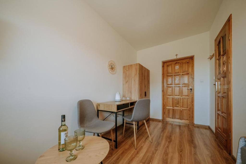 Een kleine kamer met een houten bureau, twee stoelen, een ronde tafel met glazen en een fles, en een houten garderobe met een kruis bovenop.