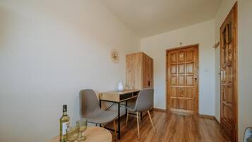 Een kleine kamer met een houten bureau, twee stoelen, een ronde tafel met glazen en een fles, en een houten garderobe met een kruis bovenop.