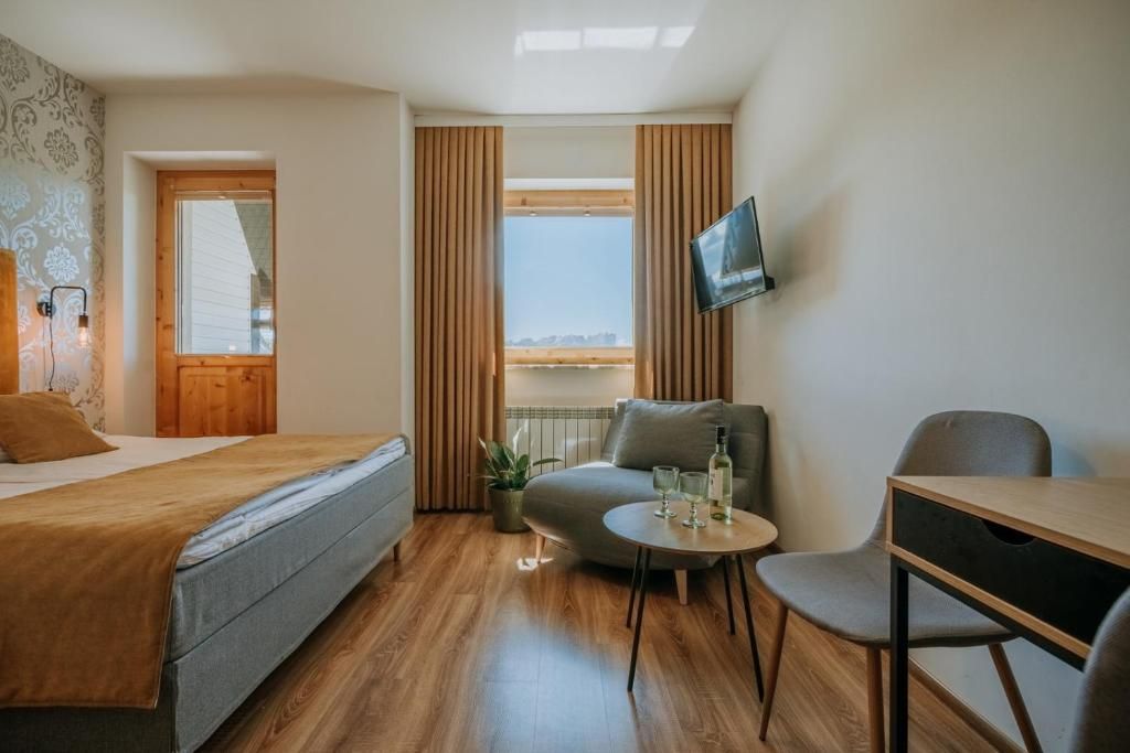 A compact hotel room with a bed, a small seating area with a table and chairs, a television mounted on the wall, and a window with curtains.