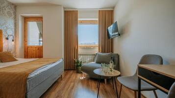 A compact hotel room with a bed, a small seating area with a table and chairs, a television mounted on the wall, and a window with curtains.