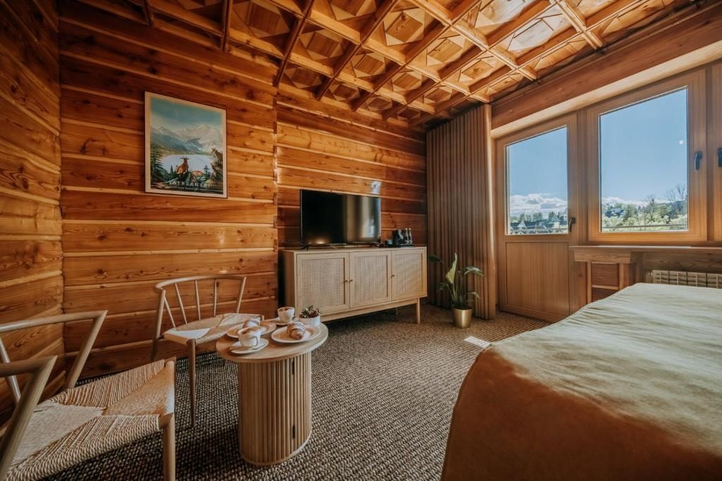 A wooden cabin room with a bed, a TV, a dining table with plates, and a window with a view.