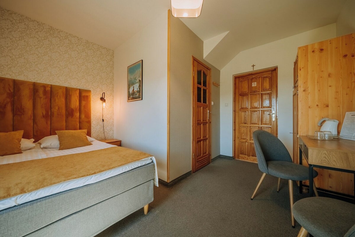 A hotel room with a bed, a desk with a chair, a wooden wardrobe, and a door with a cross.