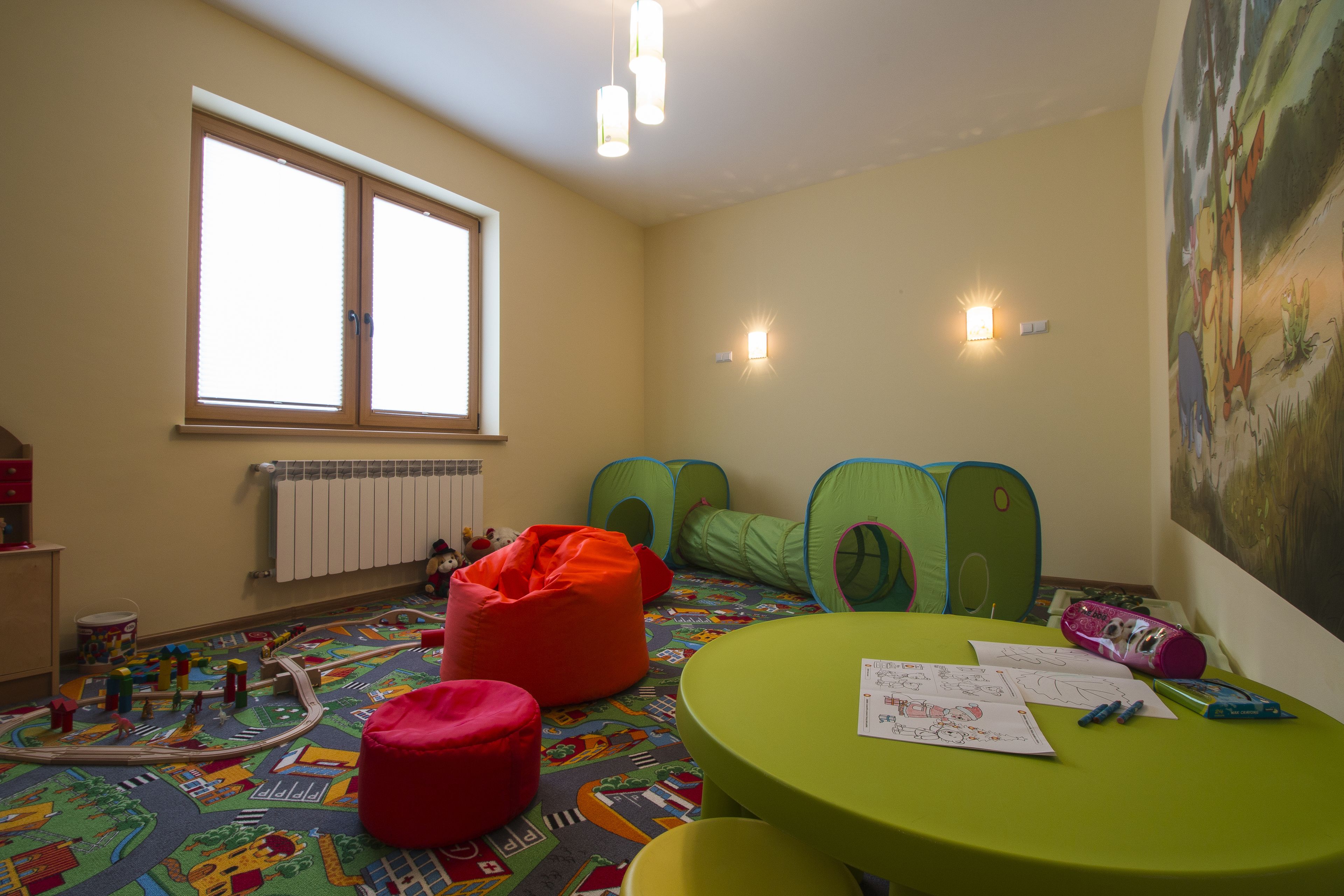 Un cuarto de juegos infantil con un tapete de colores, pufs y una mesa verde con papeles y marcadores.