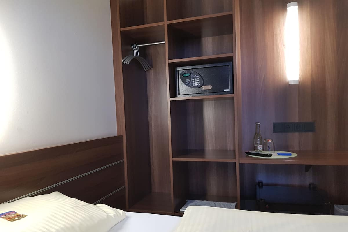 A hotel room with a bed, a wooden wardrobe, a minibar, and a bedside table with a bottle and a remote control.