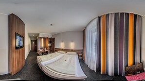 Quarto de hotel com cama curva, televisão na parede e uma grande janela com cortinas.