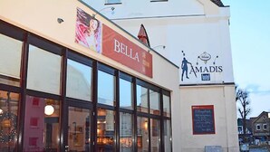 Un edificio con un letrero 'Bella' y un letrero 'Amadis', que cuenta con grandes ventanales y un interior visible con mesas y sillas.