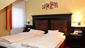 Una cama doble con sábanas blancas, cabecera de madera oscura, mesita de noche con lámpara y teléfono fijo en la pared.