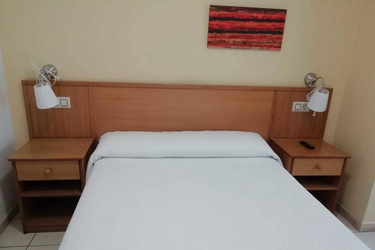 A hotel room with a wooden headboard, a white bedspread, two bedside tables with lamps, and a red abstract painting on the wall.