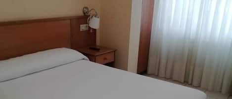 Habitación de hotel con cama, mesita de noche, lámpara, un cuadro en la pared y una ventana con cortinas.