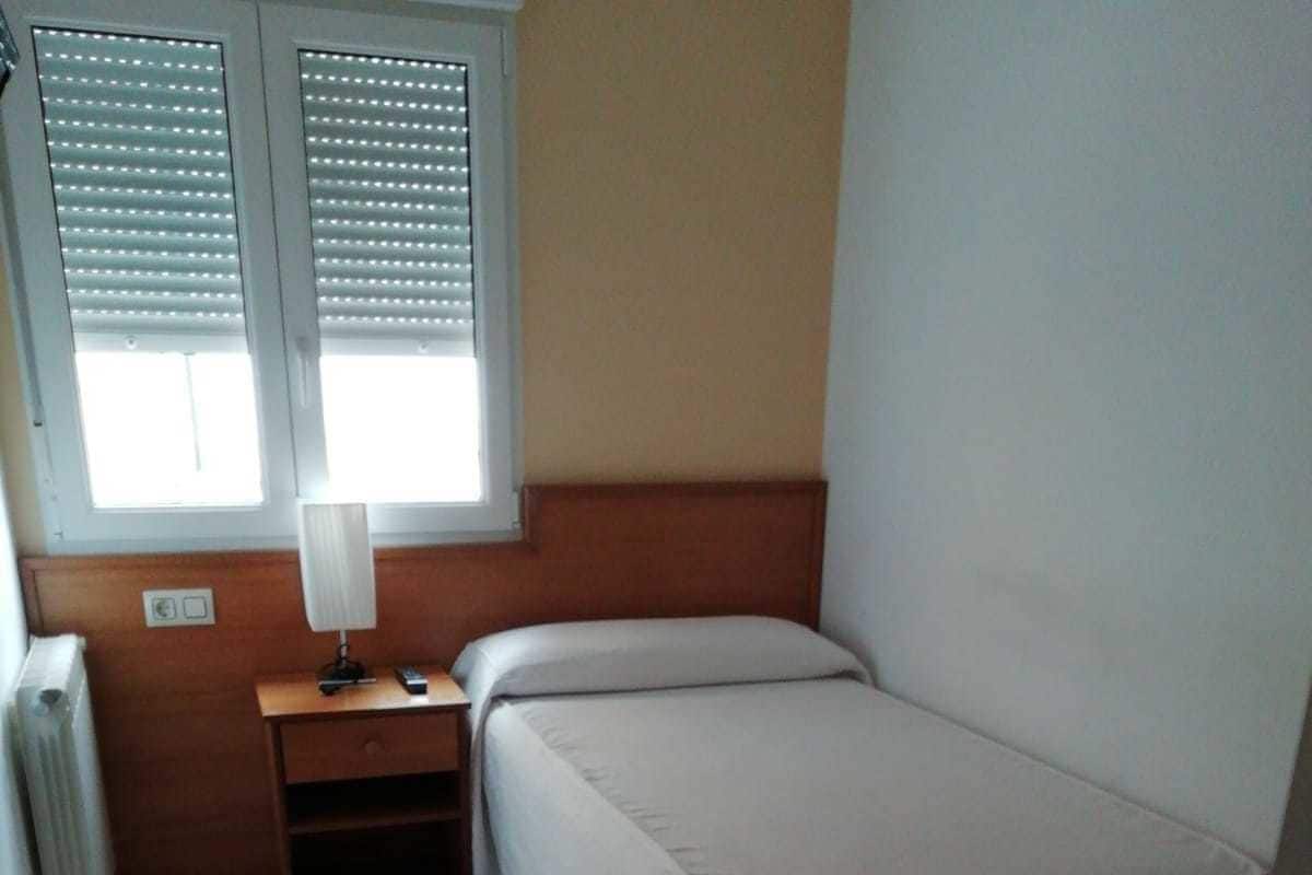 A small, clean room with a bed, a nightstand, a lamp, and a window with blinds.