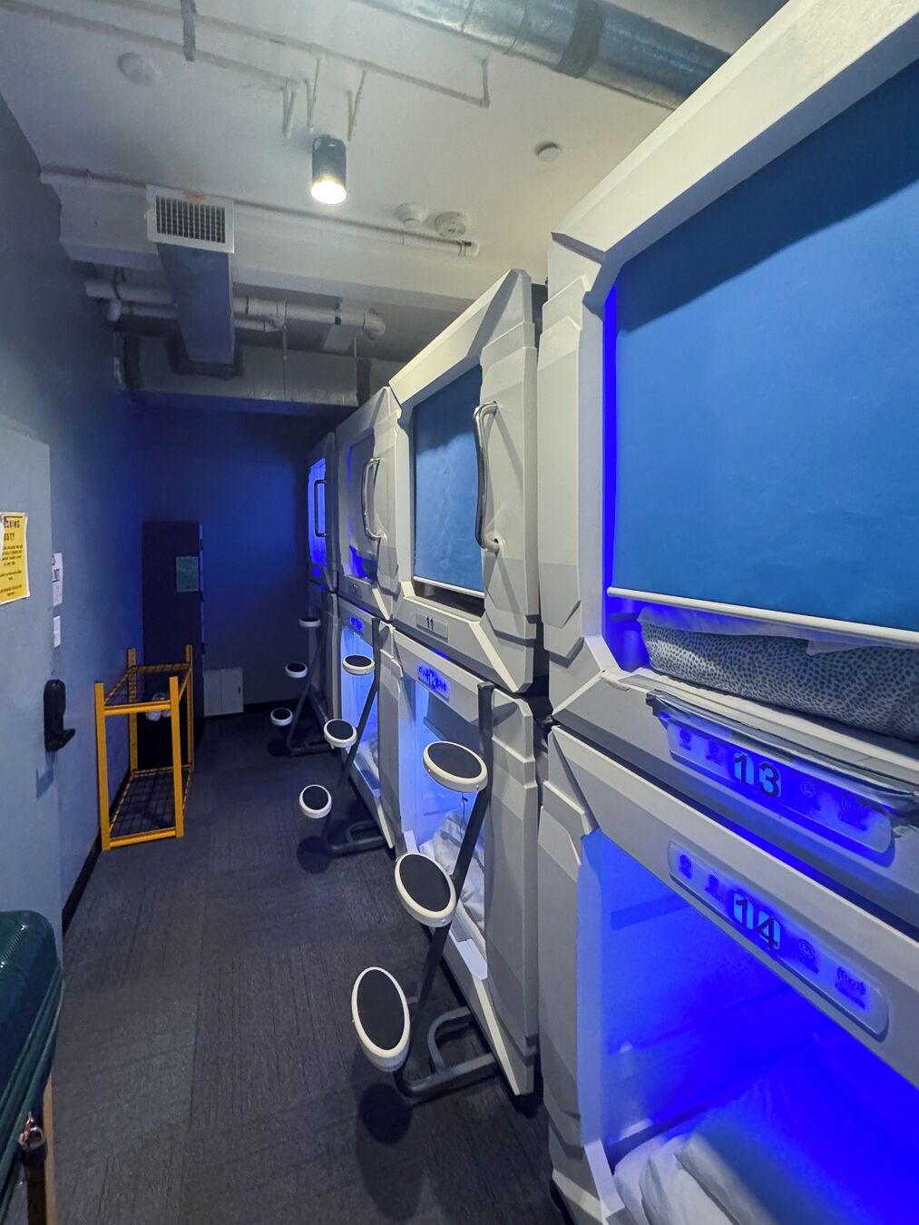 A row of compact, modern-looking rooms with individual control panels, a visible door, and a small table with a chair.