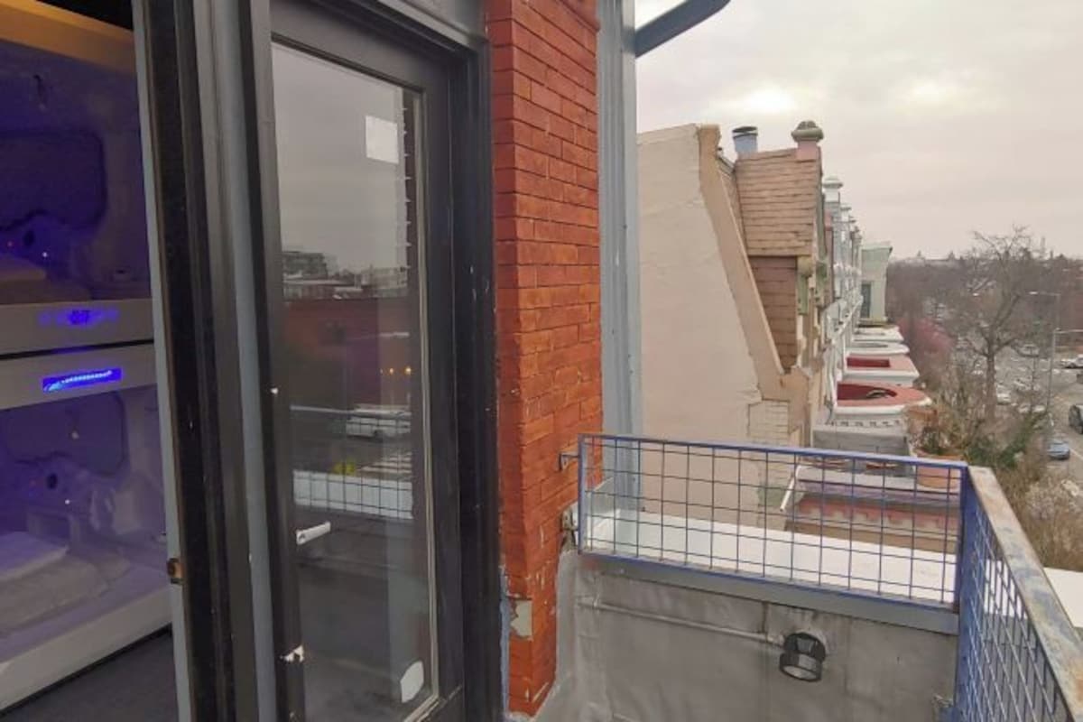 Un balcón con barandilla metálica, puerta de vidrio y vista a edificios vecinos.