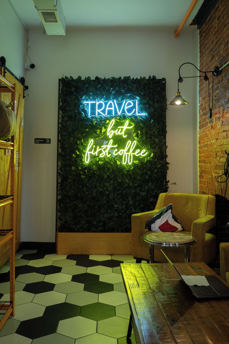 Una habitación con un letrero de neón que dice 'TRAVEL but first coffee', un banco de madera, un sillón amarillo y un suelo con patrón hexagonal.