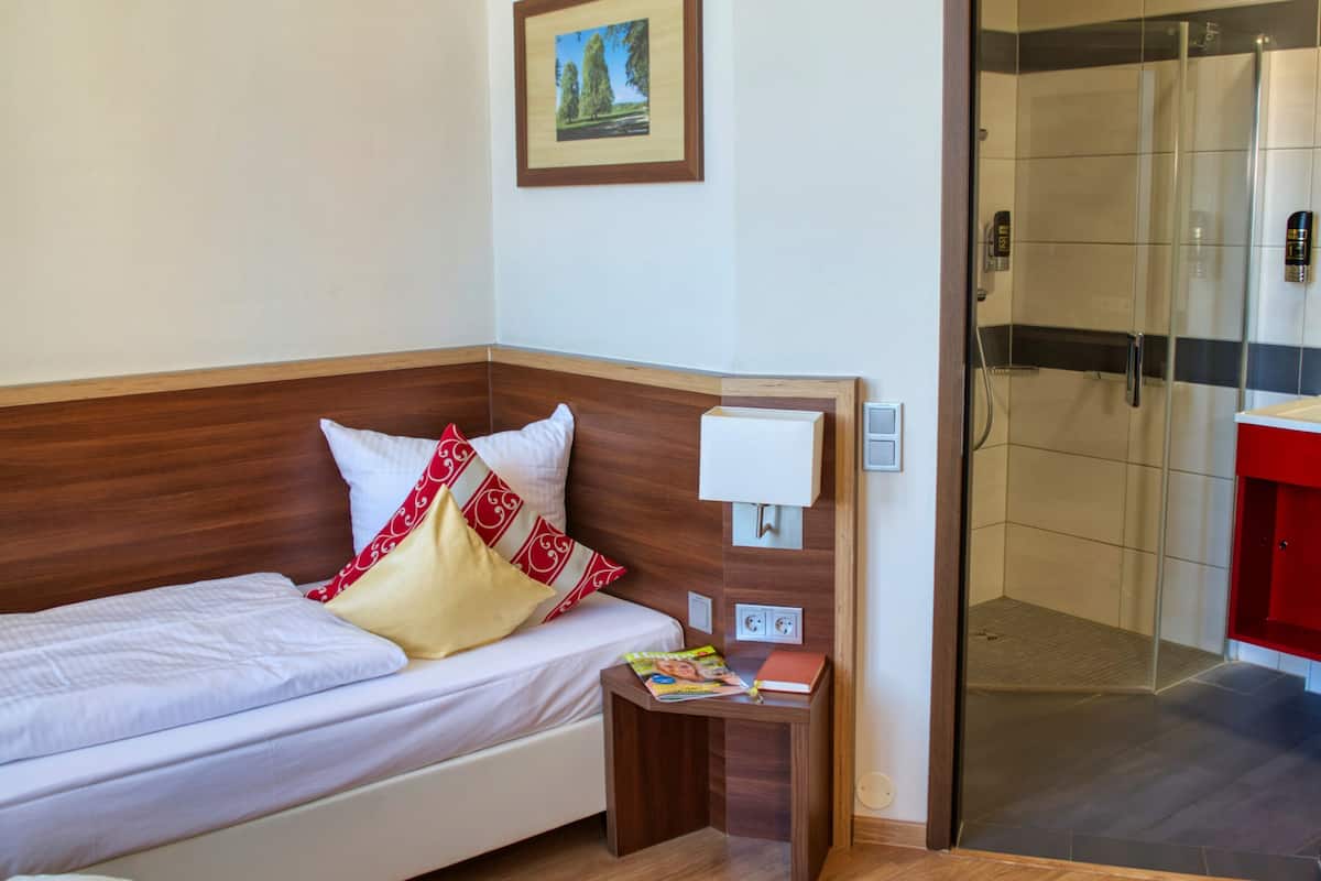 Quarto de hotel com cabeceira de madeira, cama com roupa de cama branca, almofadas vermelhas e brancas, mesa de cabeceira com revista, luminária fixada na parede e divisória de chuveiro em vidro.