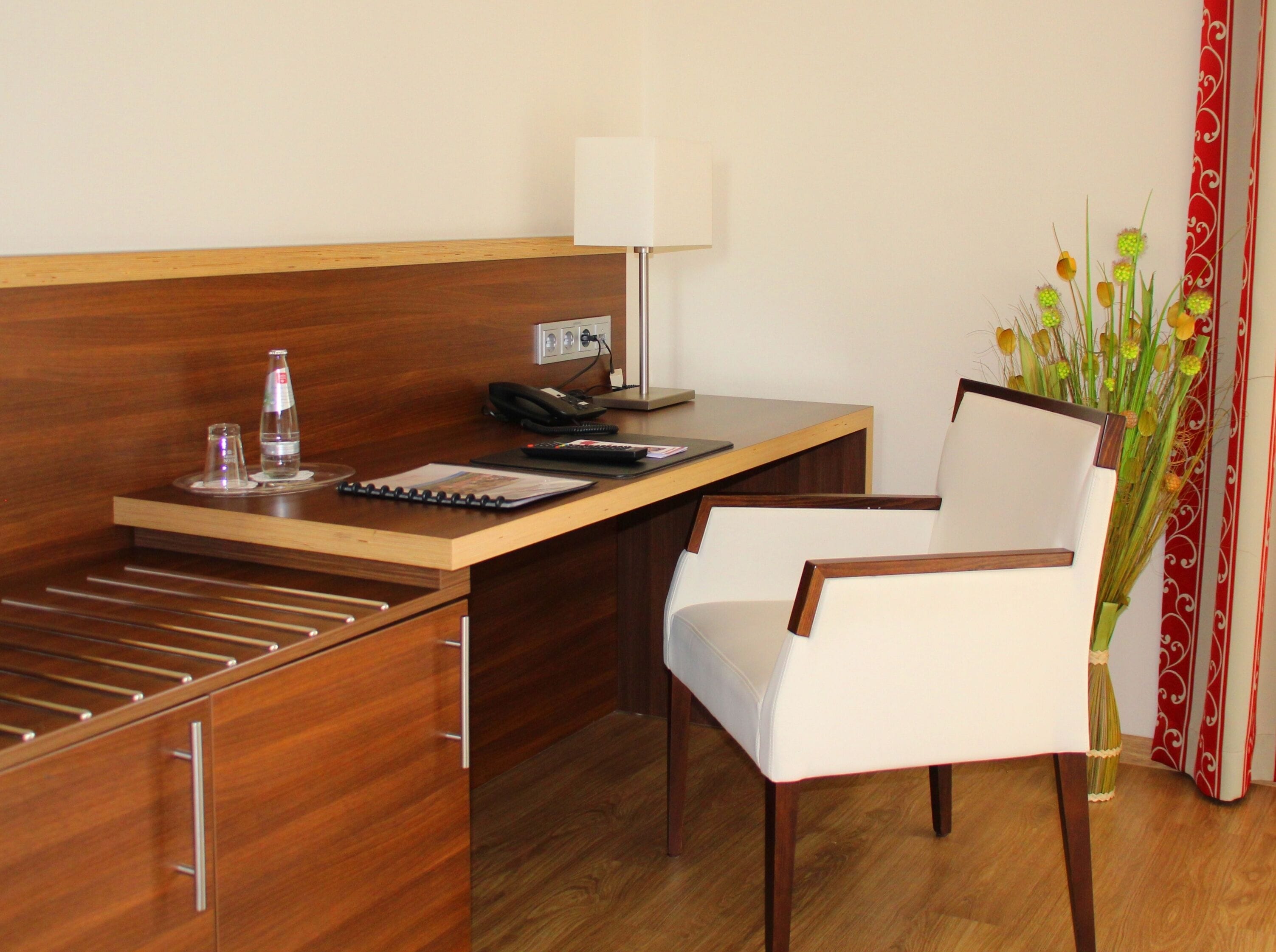 A hotel room with a wooden desk, a chair, a lamp, a phone, a water bottle, and a vase with flowers.