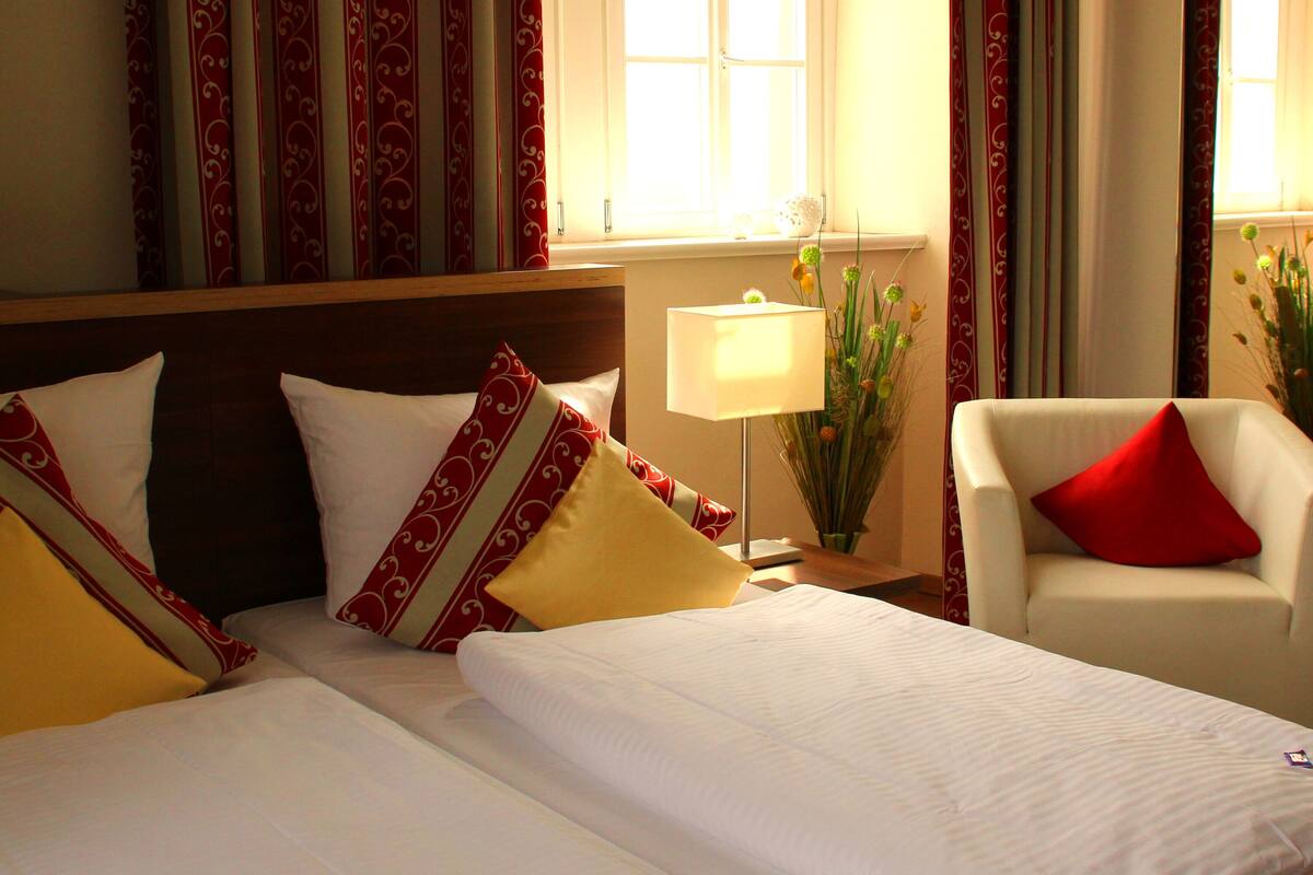 Quarto de hotel com duas camas, uma mesa de cabeceira com abajur, uma cadeira e uma janela com cortinas.