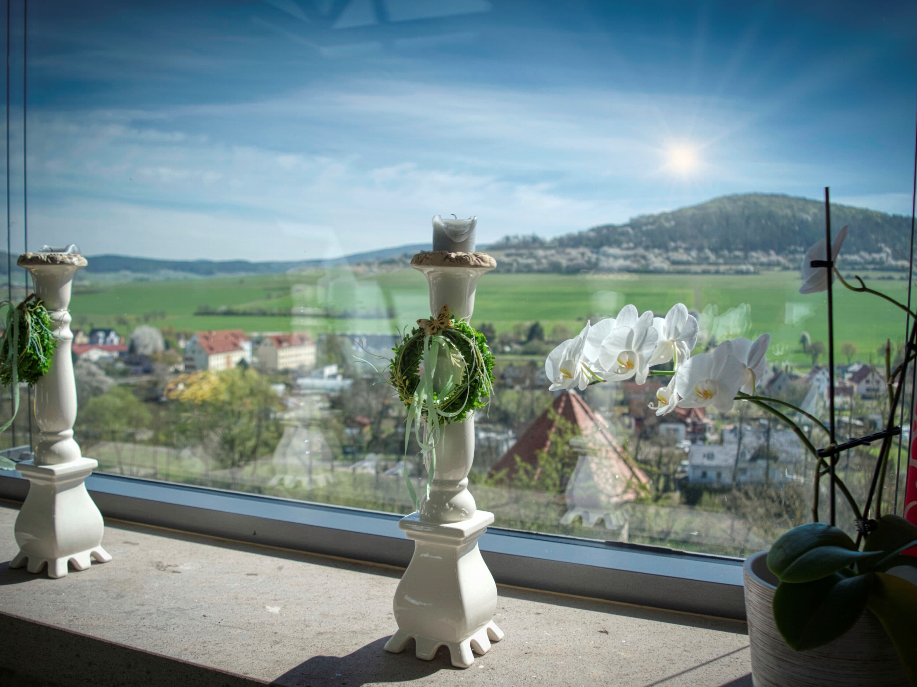 Una ventana con dos candelabros blancos decorados con follaje, que da a una vista panorámica de un pueblo y campos verdes.