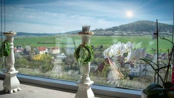 Una ventana con dos candelabros blancos decorados con follaje, que da a una vista panorámica de un pueblo y campos verdes.