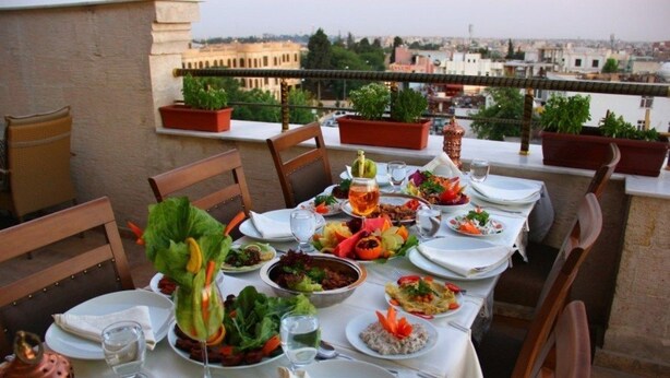 A rooftop dining table set with various dishes and drinks, surrounded by potted plants and overlooking a cityscape. - Kasr-ı Sercehan Hotel (Nusaybin)