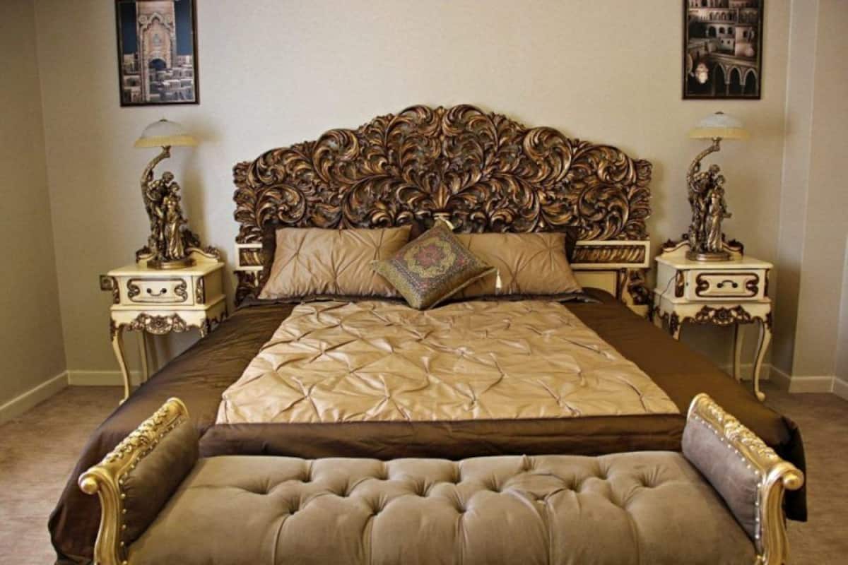 A luxurious bedroom with an ornate headboard, a tufted bench, and decorative lamps.